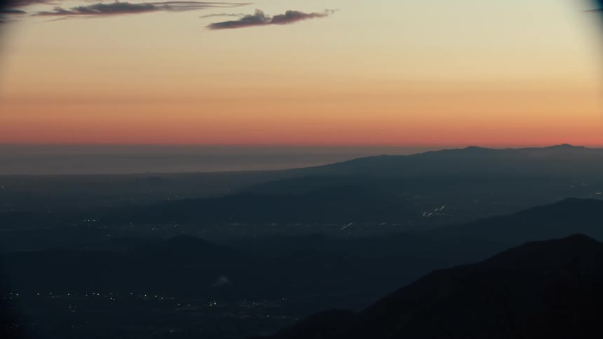 Mountain vista during twilight where the sky is dominated by a warm gradient of deep orange and gold silhouetting layers of dark distant ridges that stretch toward the horizon beneath which the lights of a settlement are just visible