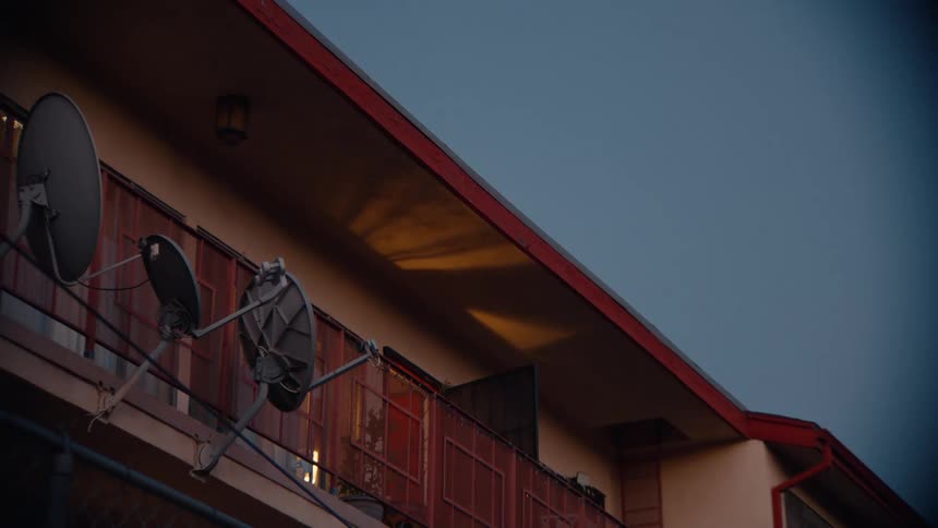At dusk a slow pan reveals the balconies of a weathered apartment building featuring satellite dishes and fading light against a deep blue sky