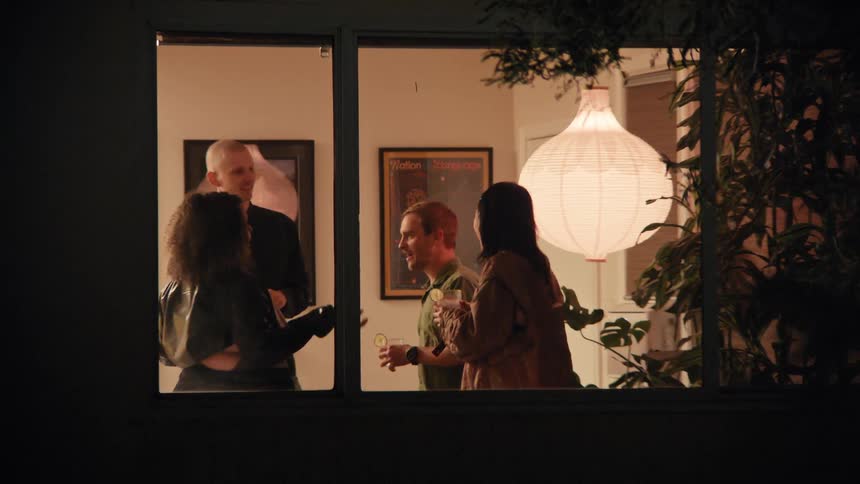 Four friends are drinking and chatting happily as seen through a window one night