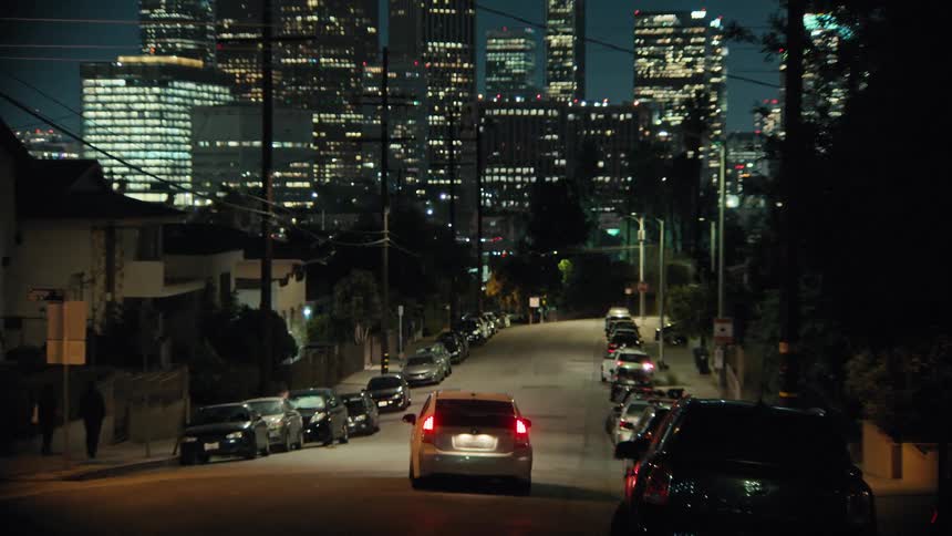 People get into a car then the vehicle drives down the residential street towards the city at night