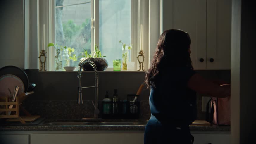 A kitchen counter and sink area fully illuminated by bright sunlight streaming through a large window silhouetting a woman with long dark hair as she finishes placing sandwiches in a bag and then walking away