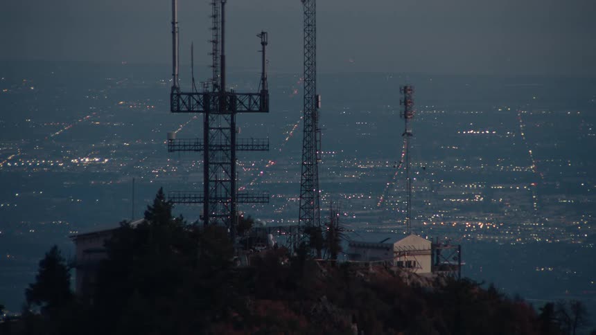 A group of steel communication towers and accompanying structures silhouetted against a pale twilight sky atop a dark mountain ridge with a city below