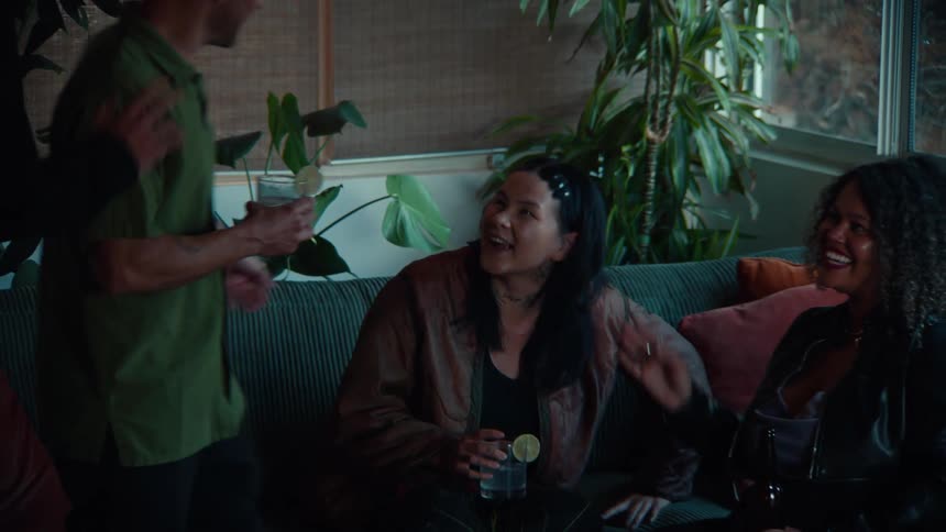 A smiling woman holding a drink then gradually zooms out to reveal her sitting on a ribbed couch alongside a diverse group of three other adults all of whom are laughing and engaging in casual conversation in a warmly lit plantfilled indoor space
