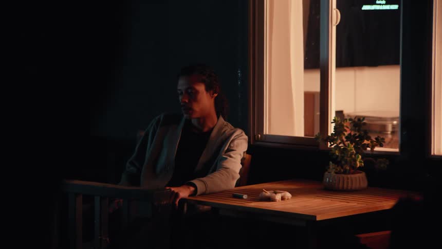 Man sits at a table on the patio outside of a house as he smokes a cigarette and thinks as another person stands nearby