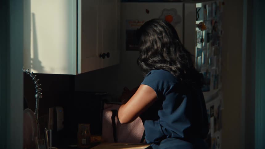 A woman with long dark hair dressed in a dark blue outfit standing in a sundrenched section of her kitchen as she meticulously places a sandwich or similar food item into a plastic bag and then into a portable lunch bag
