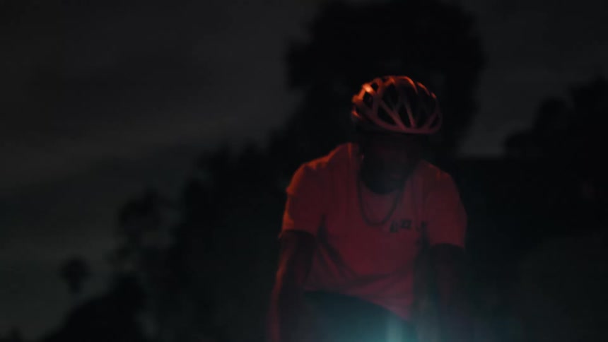 A man wearing a helmet and a lightcolored tshirt cycling directly towards the viewer on a dark residential street at night with the scene largely silhouetted and obscured by the intense blinding flare emanating from the bicyclemounted headlight
