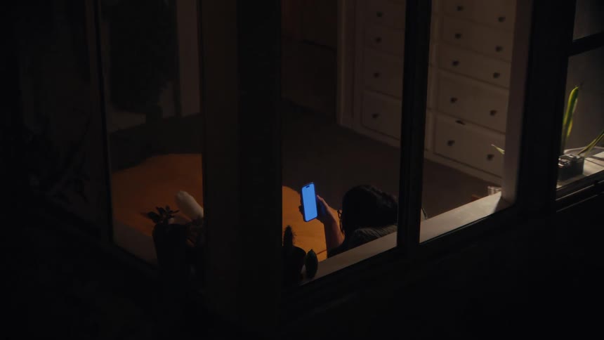 A female sits near a window of a room while looking at her smart phone