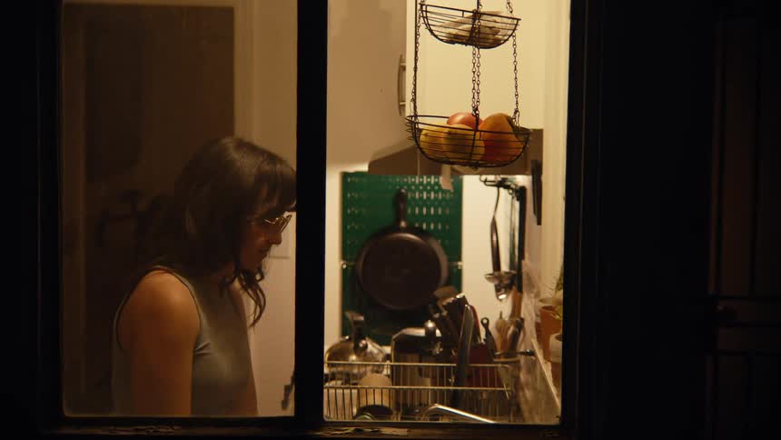 A woman with dark hair and glasses in a brightly lit kitchen at night observed from outside a window as she quietly works at the sink area handling items in a dish rack with a hanging fruit basket and various kitchen tools visible around her