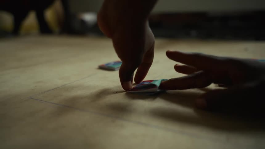 A person is flicking small colorful tiles on a wooden surface as a hand tries to block the tile movement