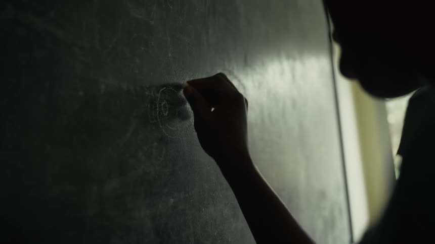 Silhouette of a person drawing with chalk on a blackboard with another person standing next to them indoors during day time