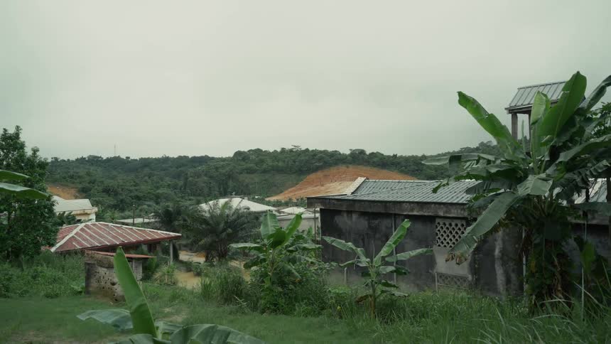 Lush tropical vegetation surrounds a small village of simple buildings nestled in the hills under a hazy overcast sky