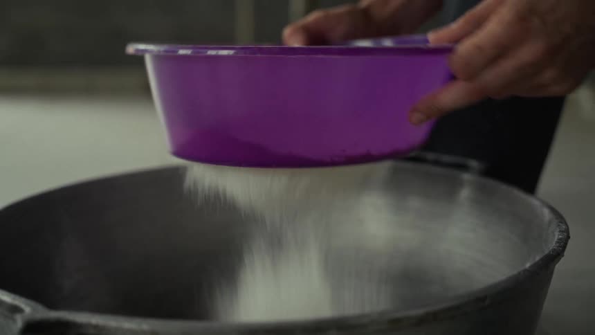 A person is pouring a stream of fine white powder likely flour or sugar from a purple plastic bowl the ingredients are being added to a large dark metal pot or mixing vessel