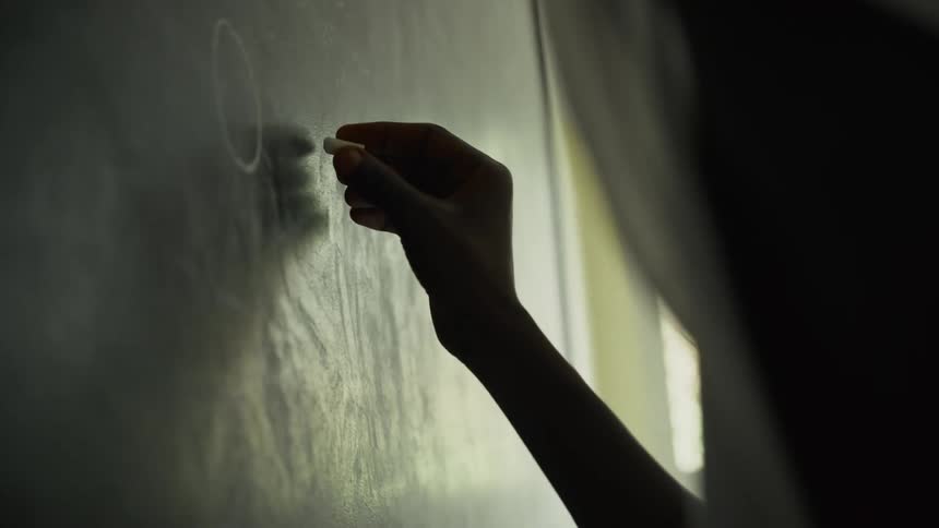 Hand writes with chalk on a board under soft light