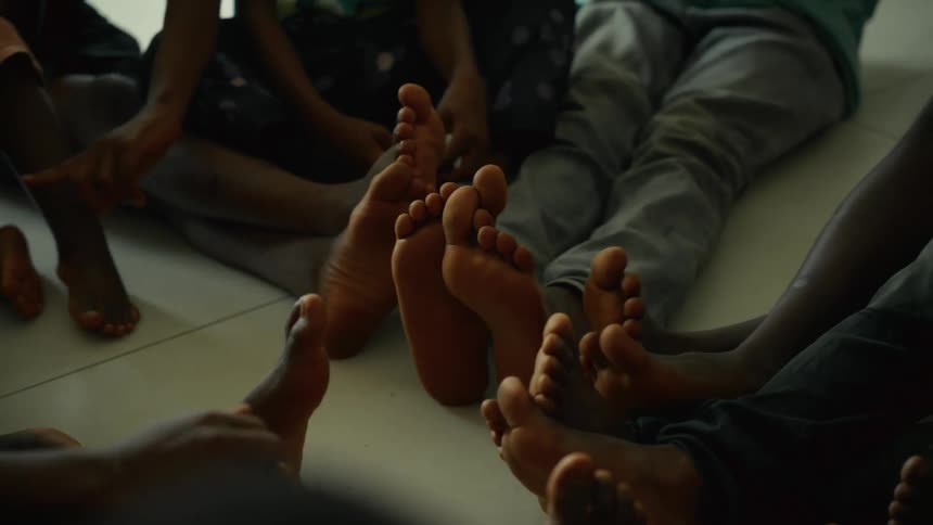 A cluster of multiple bare feet and lower legs of several individuals sitting in a close circle on a pale floor with some hands visible near the edge of the frame