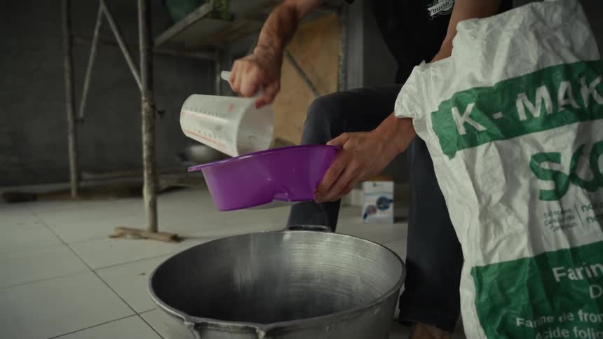 A person carefully sifts flour from a measuring cup through a purple sieve into a large metal pot with a large bag of flour leaning against them
