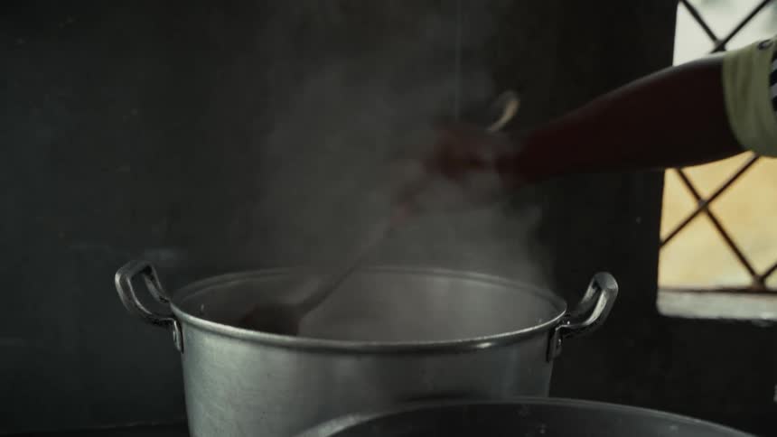 A large steaming aluminum pot in a dimly lit kitchen as a person visible only by their arm and sleeve vigorously stirs the unseen contents inside with a large metal utensil