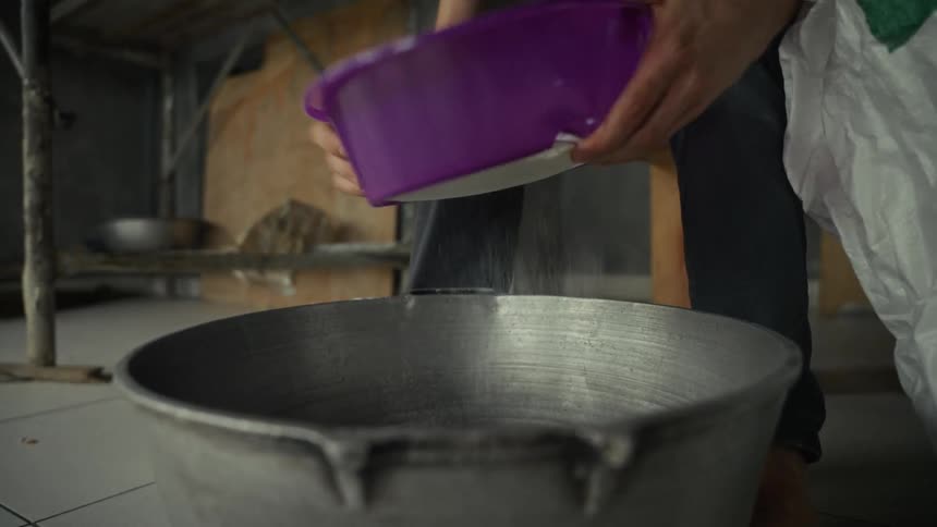 Hands pour white grains from a purple bowl into a large metal pot