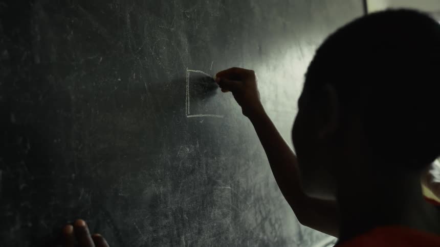 A childs silhouette is visible in the foreground as they draw a shape on a large dark chalkboard they are holding a piece of chalk and actively sketching a square or rectangular figure