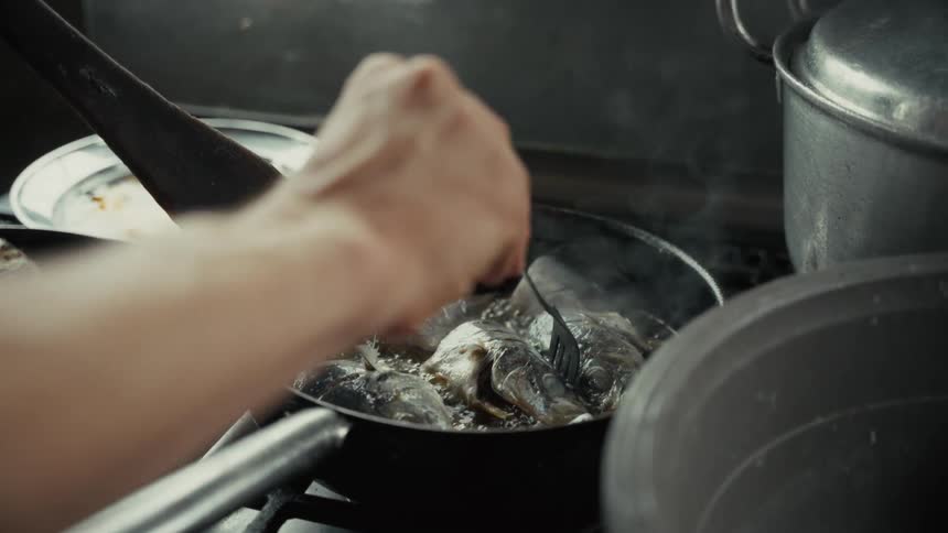 A cook uses a utensil to stir pieces of fish as they sizzle and steam in a hot frying pan