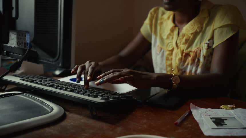 In a softly lit room a woman with colorful nail polish types on a computer keyboard at a wooden desk