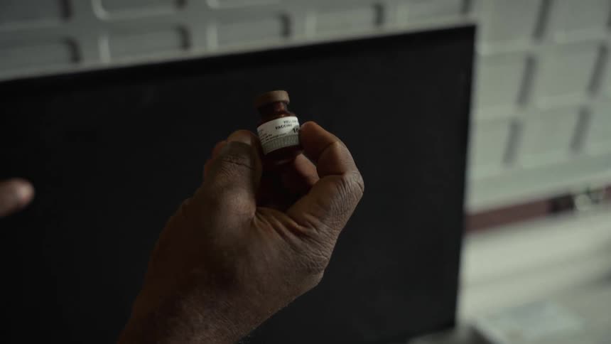 The video is a closeup shot focused on a darkskinned hand holding a small brown glass vial likely containing a medical solution or vaccinethe label on the vial is partially visible and the words yellow fever and 10 doses can be faintly read s