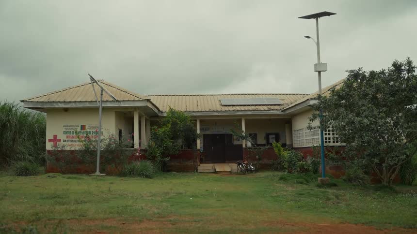The video shows a wide static shot of a singlestory modest building likely a rural health center the building has a lightcolored exterior with a reddishbrown base and a corrugated metal roof and importantly it is equipped with solar panels mount