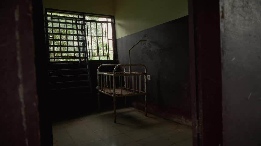 A dimly lit empty room with a metal crib in the center standing near a barred window letting in greentinged light