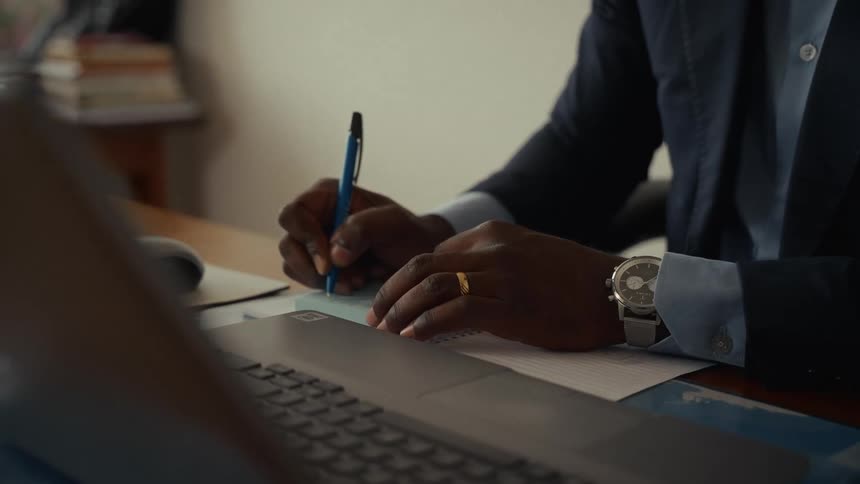 Man sitting at a desk holding a computer writes on a piece of paper