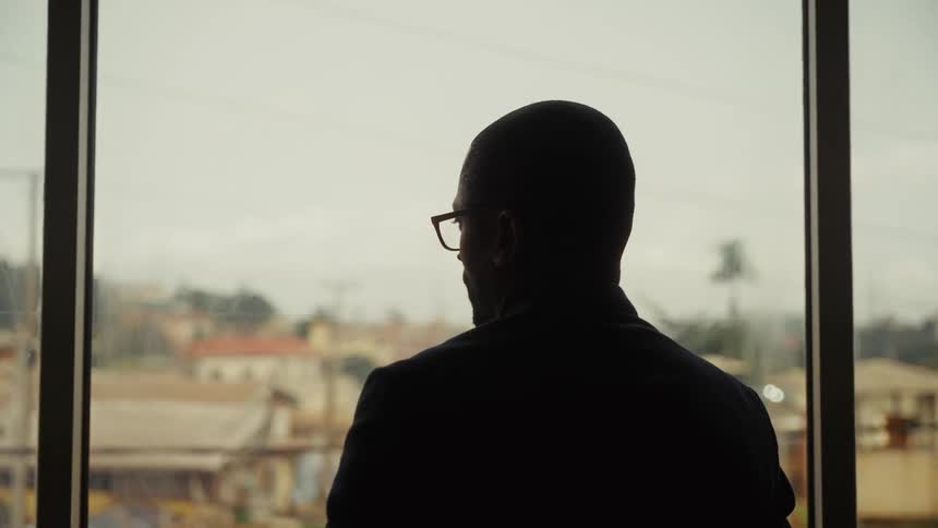 A silhouetted man wearing glasses stands before a large window gazing out over a hazy cityscape softly lit by muted daylight