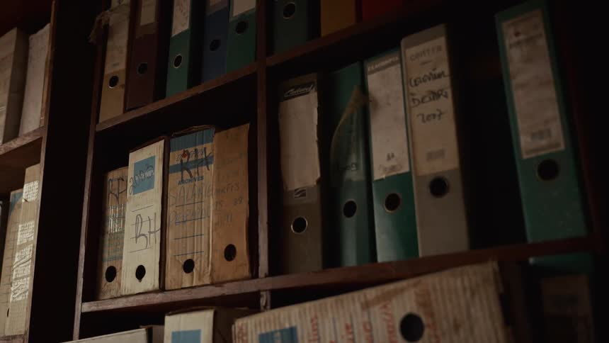 A dimly lit wooden bookshelf filled with assorted binders in various colors including beige blue green and white the binders have labels and holes with some showing signs of wear