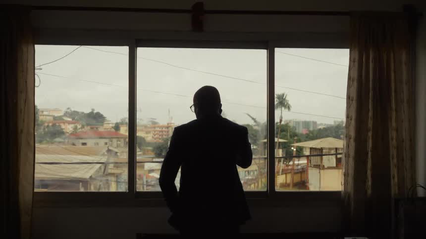 The video shows a view of a densely built hilly urban landscape seen through a threepane window featuring mixeduse buildings with various roof types under an overcast skya man dressed in a suit walks into the frame and stands silhouetted agai