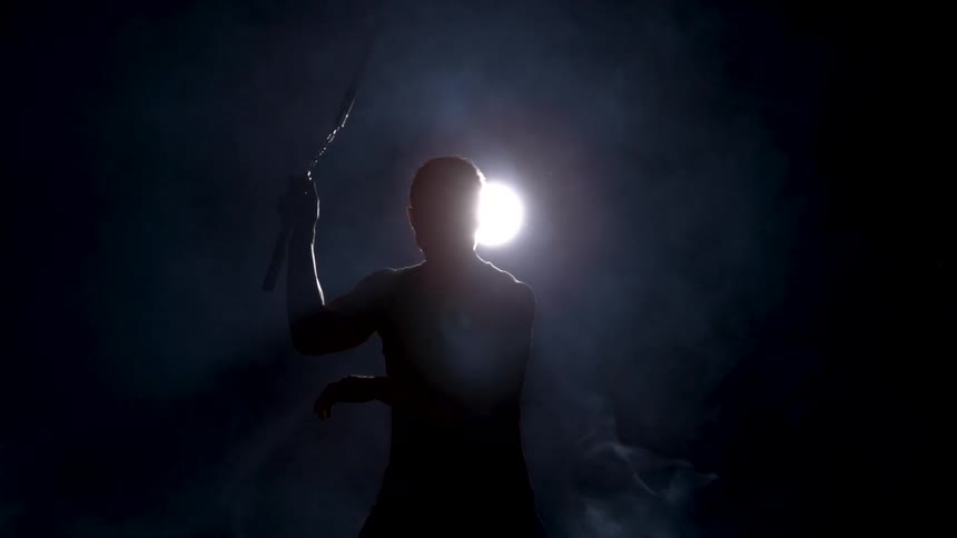 Man cast into silhouette by a spotlight practices with nunchucks