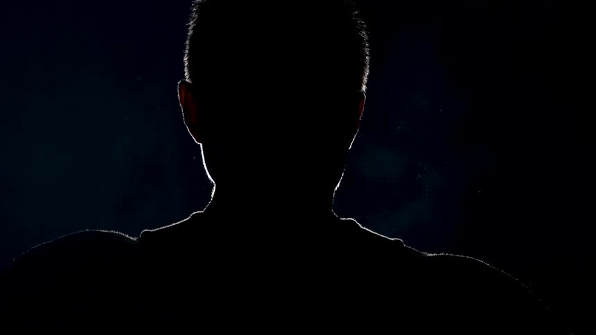 A silhouetted person with short hair partially illuminated against a dark nearly black background is using nunchucks in one hand