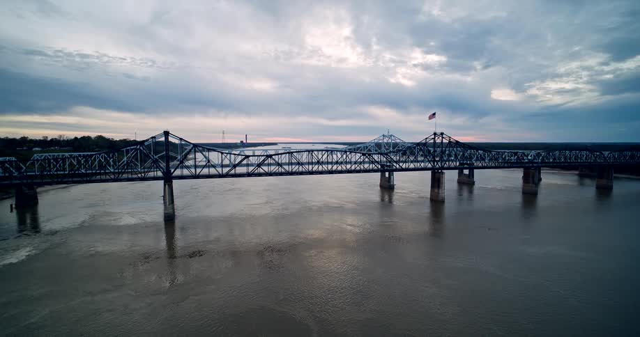 Aerial view of a steel bridge over a wide river at sunset under a cloudy sky