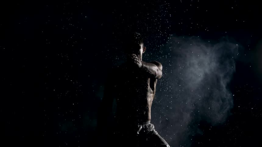The clip features a dark lowkey setting highlighting a shirtless muscular person who is moving their arms as if exercising or performing a dramatic motionwater or dust particles are raining down and mist is rising around them catching the light and