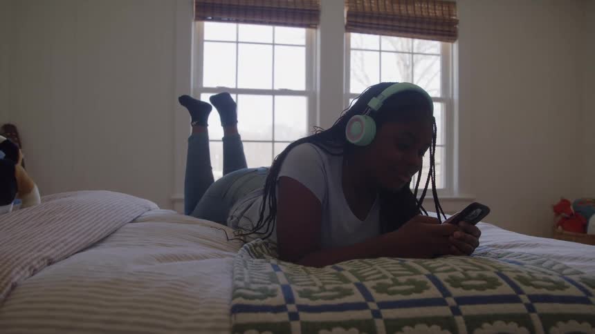 A teenage girl lies on her bed while scrolling on her smartphone