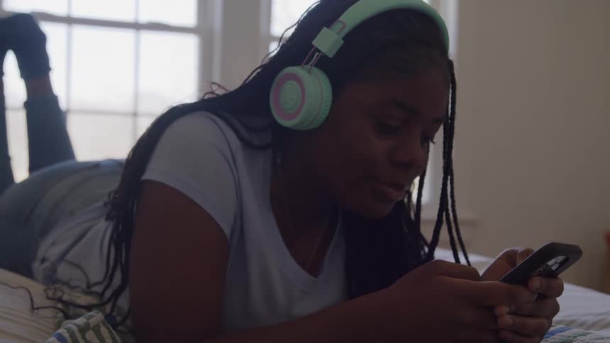 A young girl with braided hair lies on her bed listening to music with headphones while using her smartphone