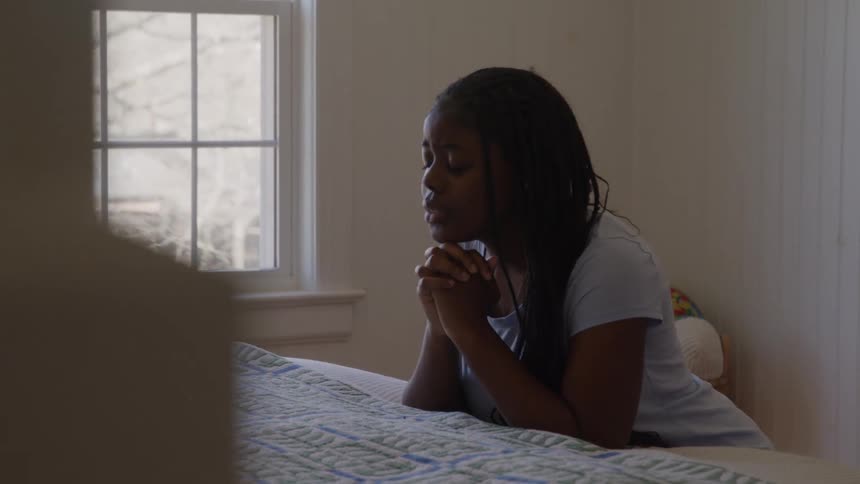 A young woman kneels at her bed while praying out loud