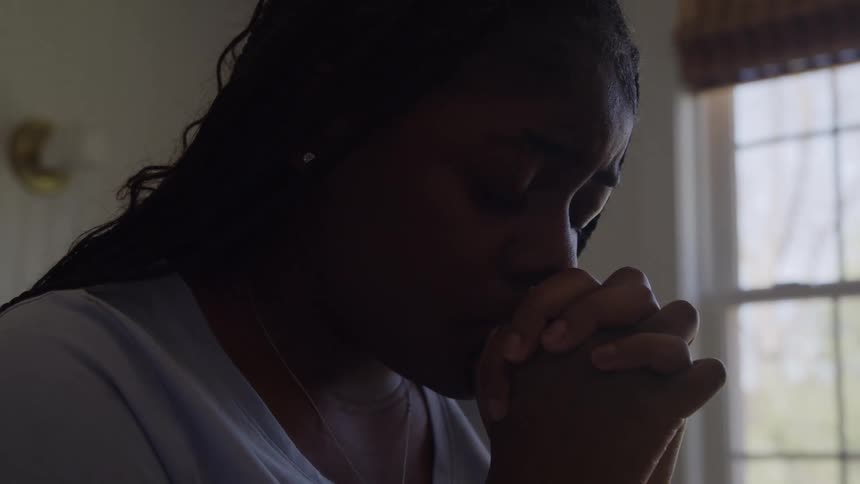 A sad woman sits indoors by a window with her hands clasped