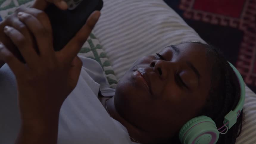 A young person is lying down wearing mint green and pink headphones while looking intently at a black smartphone held in their handsthe lighting is dim suggesting they are relaxing or watching content in a lowlight setting