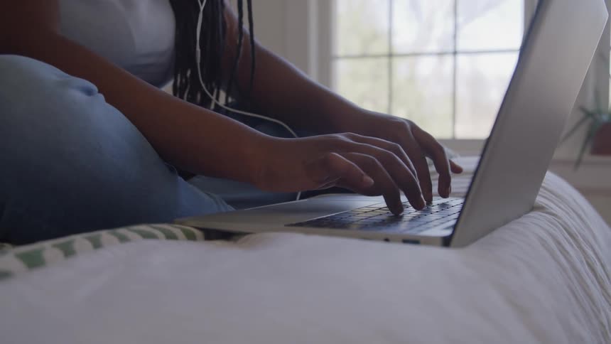 A young woman sits on her bed while typing on her laptop keyboard