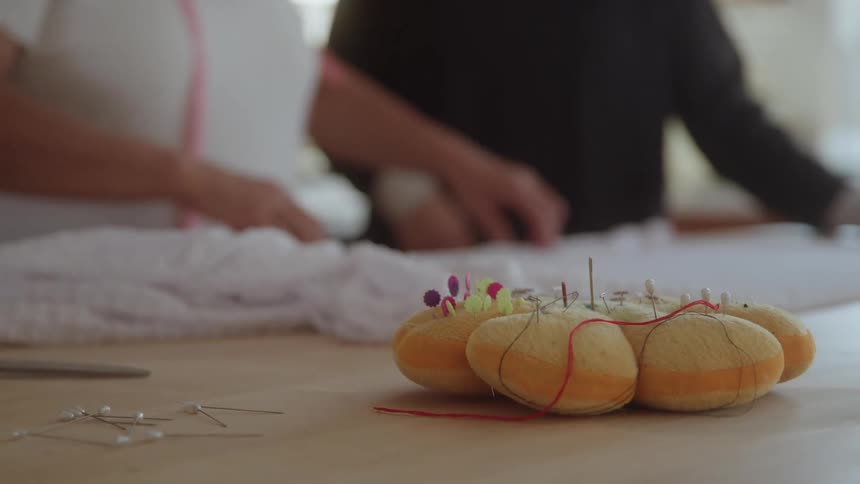 The foreground features several small orange and yellow pumpkinshaped pin cushions with various sewing pins stuck into them lying on a wooden surfacein the blurred background two people are seen working likely on a sewing or fabric project with