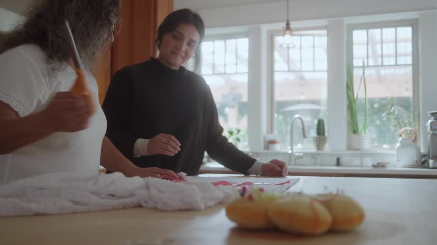 In a sunlit kitchen an older woman teaches a younger woman how to cut fabric for a sewing project at the kitchen island