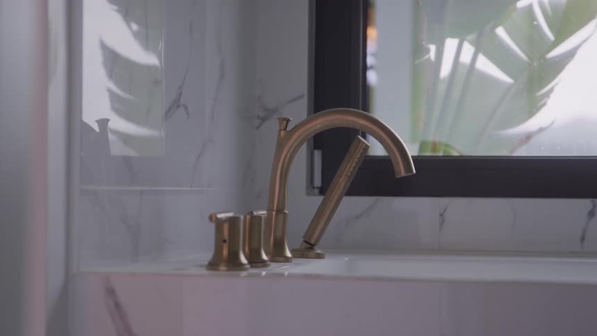 A modern brass faucet is elegantly mounted on the edge of a bathtub set against a white marble wall next to a large window with plants visible outside