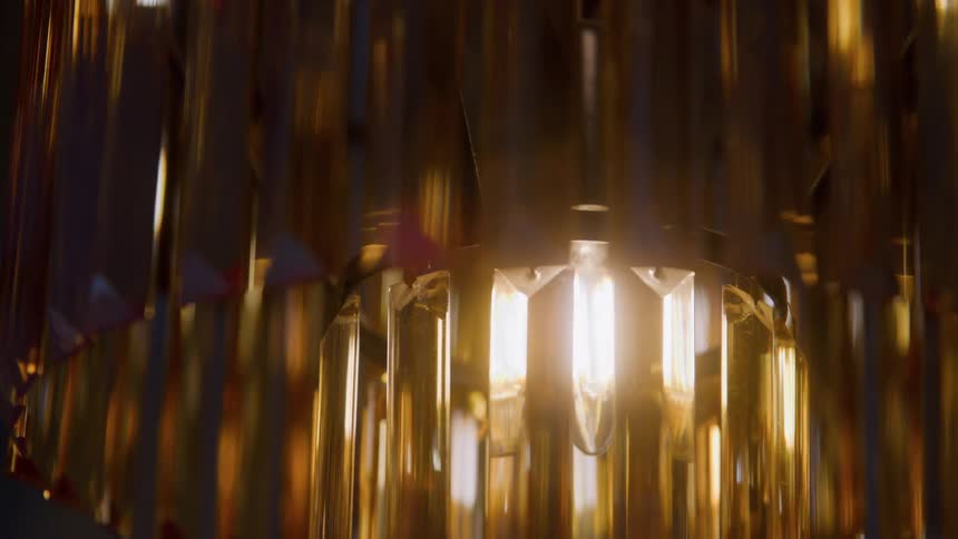 The center of a chandelier capturing a single brightly lit incandescent bulb and its warm glowing light shining through and being refracted by surrounding vertical rods made of golden material