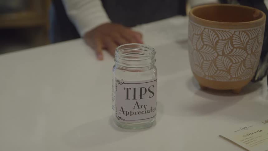 A customers hand places a folded cash tip into a glass jar labeled tips are appreciated sitting on a white cafe counter