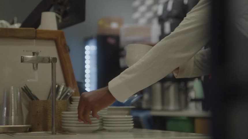 The service counter of a coffee shop where a barista dressed in a white shirt and dark apron hands a white cup and saucer to a costumer