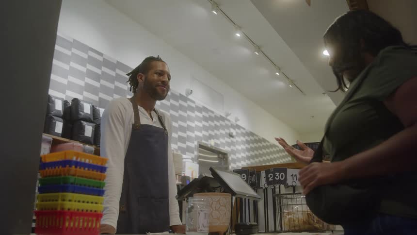 A male barista wearing a white shirt and a dark apron is standing behind a counter and interacting with a female customer on the rightthe two people appear to be engaged in a pleasant conversation possibly as the customer is placing an order or payi