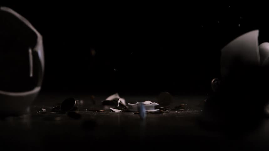 A ceramic white piggy bank is dropped from above onto a dark surface shattering immediately upon impactcoins of various denominations scatter outwards in slow motion as the pieces of the broken piggy bank fly apart against a completely black backgroun