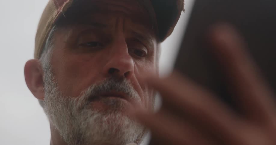 An elderly man with white facial hair wearing a hat operating his mobile device outdoors at midday with a sad expression